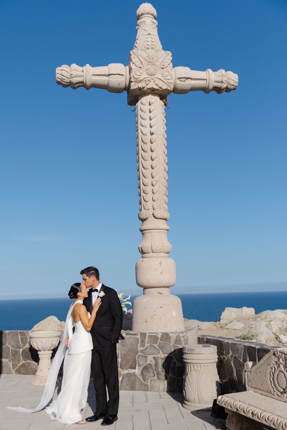 Wedding couple kissing beneath an ornate stone cross monument with clear blue sky and ocean in the background.