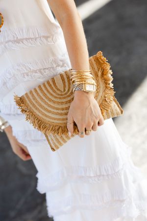 Close-up of a woman in a white ruffled dress holding a woven straw fringe clutch with gold bangles and watch.