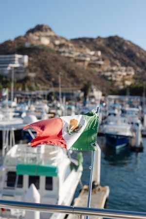 Mexican flag waving in the foreground with a marina full of boats and a rocky hillside in Cabo San Lucas.