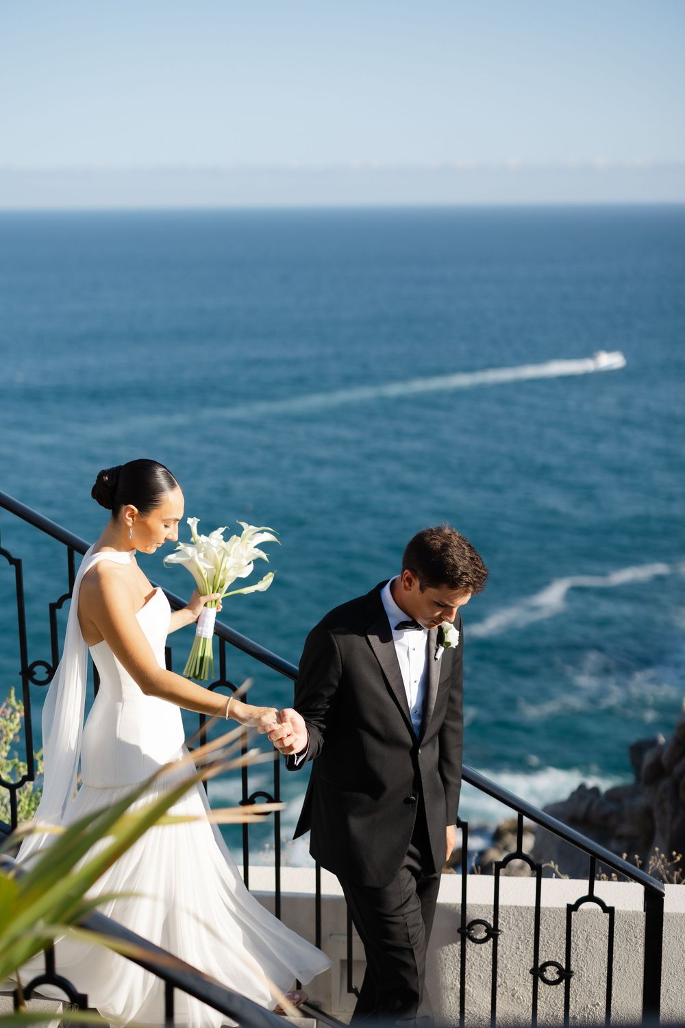 Bride and groom holding hands descending stone stairs with stunning turquoise ocean views behind them on their wedding day.