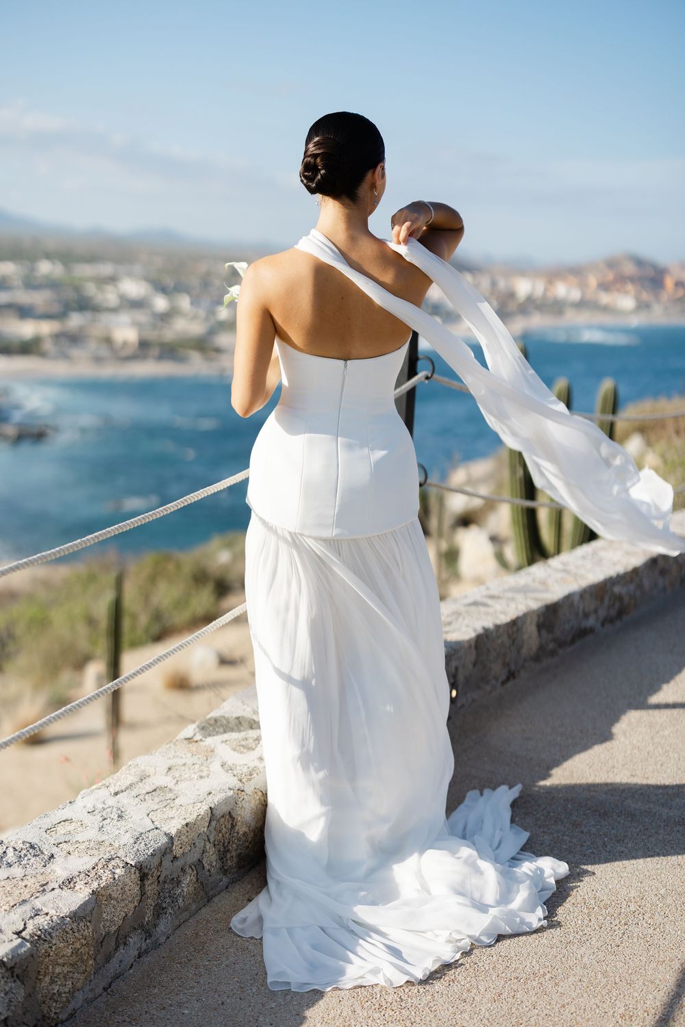 Bride in a backless white wedding gown standing on a scenic overlook with ocean and coastal town views in Cabo.