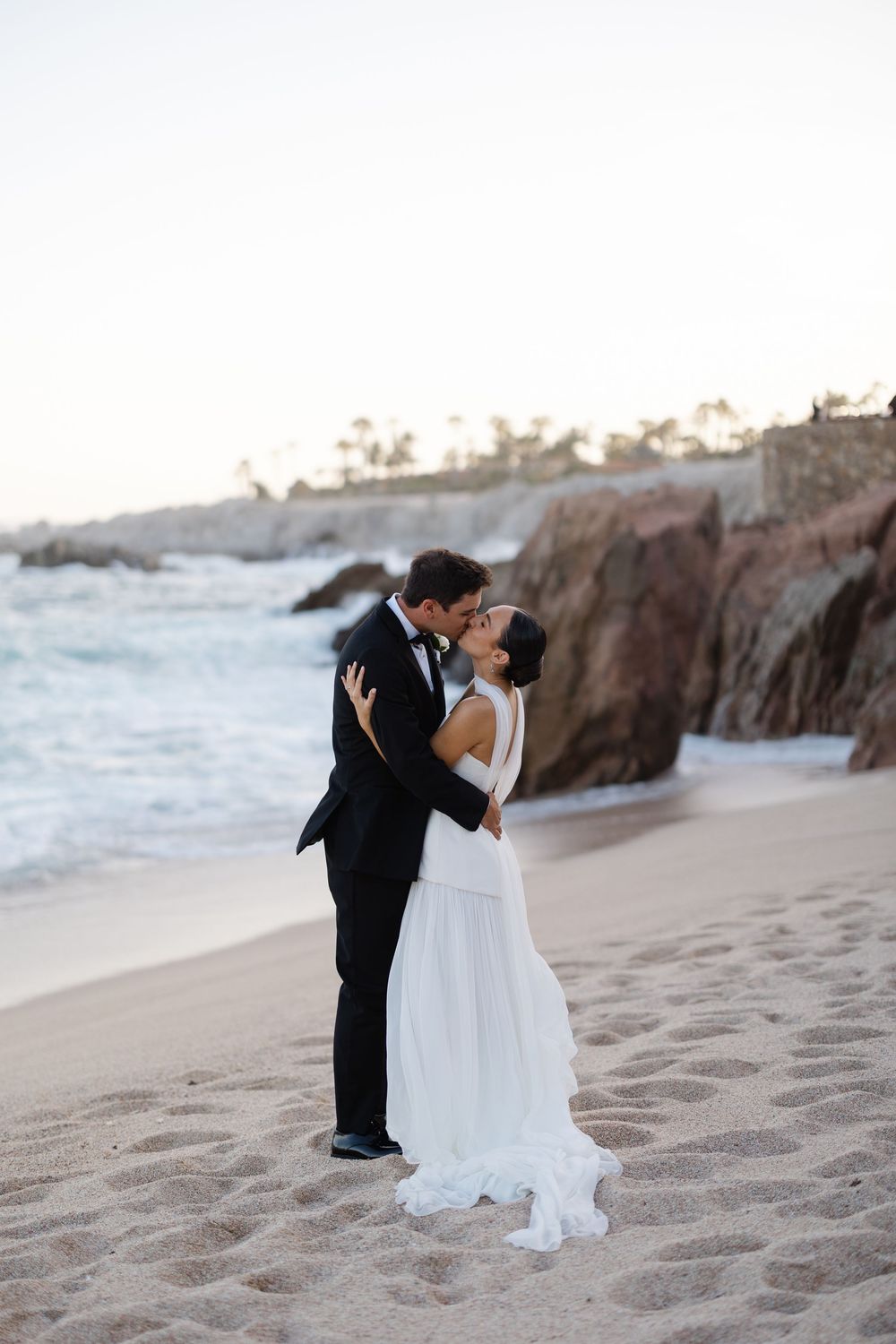Bride and groom sharing a romantic kiss on a sandy beach at dusk with rocky cliffs in the background.