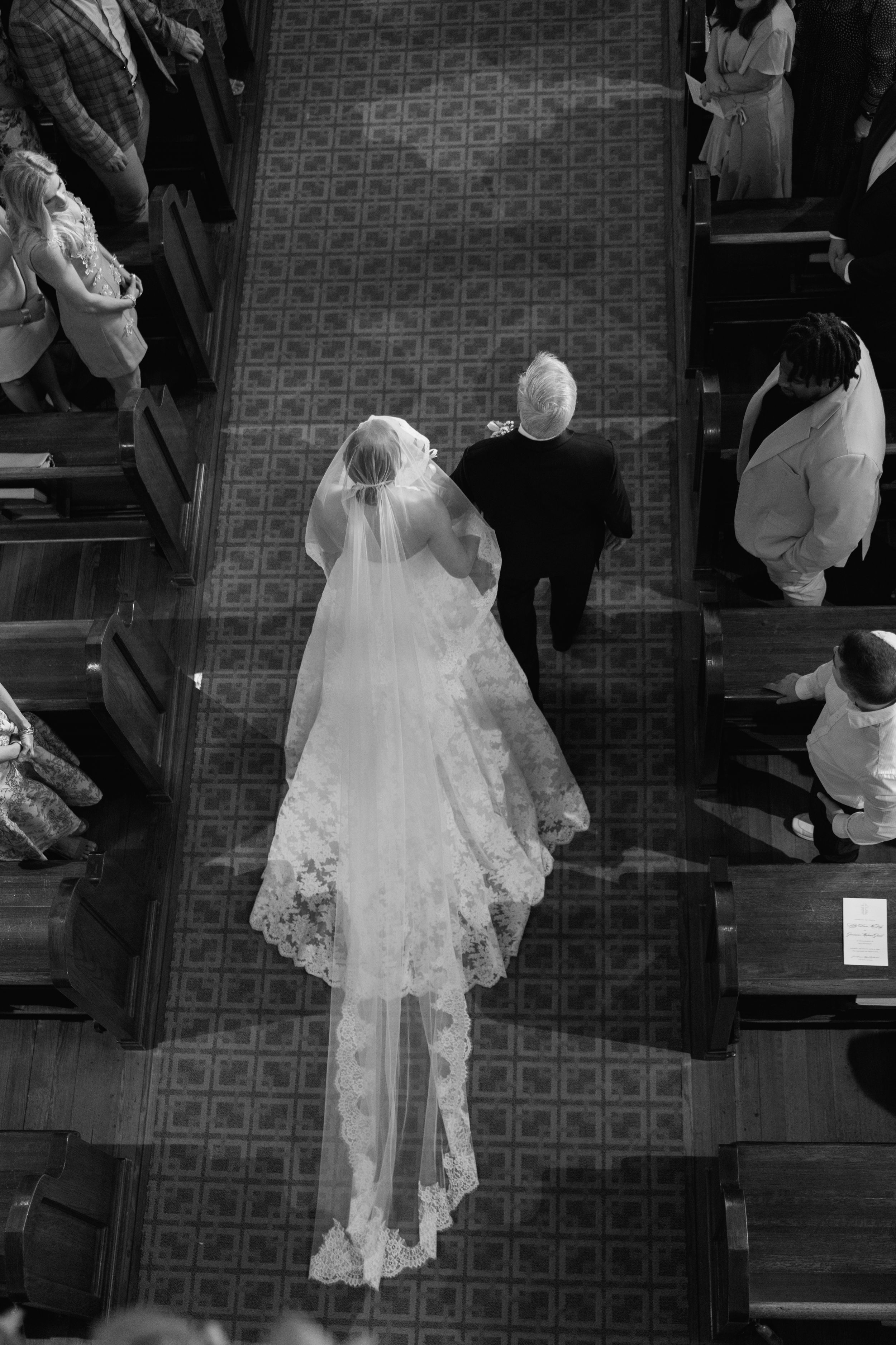 Aerial black and white view of bride walking down church aisle with long lace veil trailing behind.