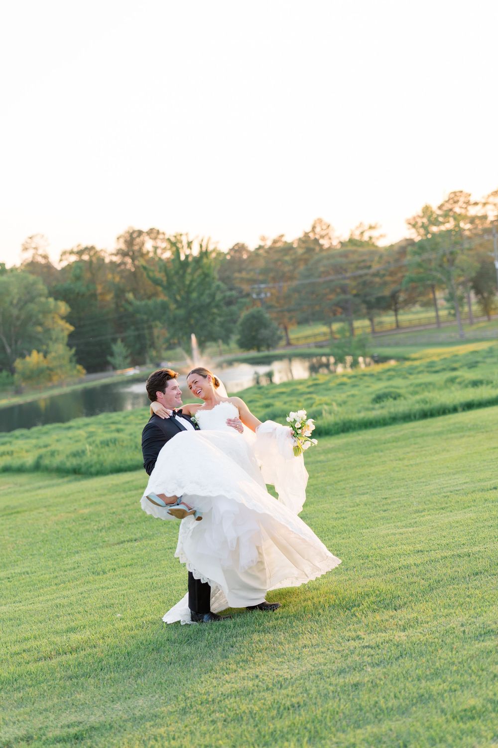 Groom lifts laughing bride in wedding gown on lush green lawn at golden hour near a pond.