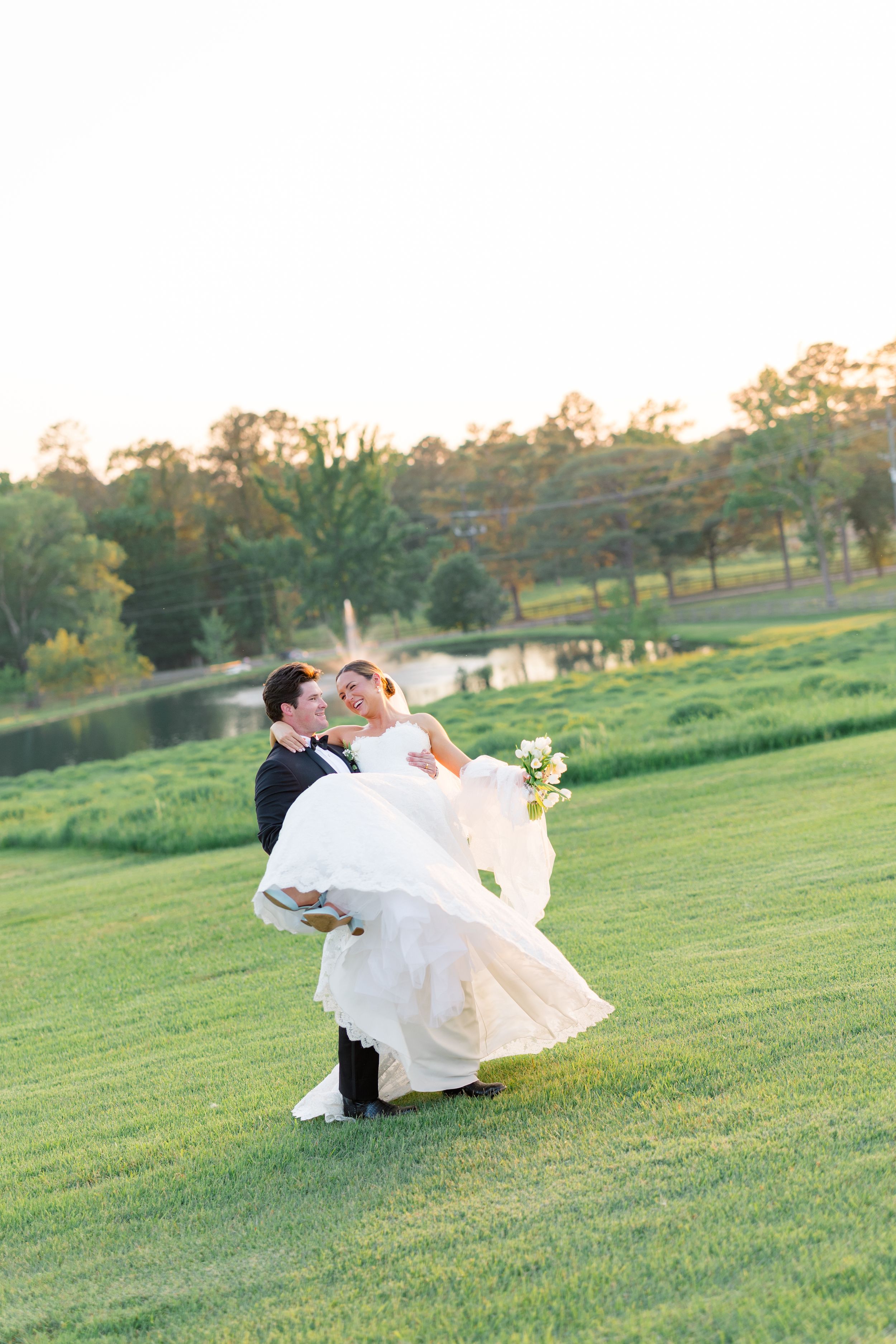 Groom lifts laughing bride in wedding gown on lush green lawn at golden hour near a pond.