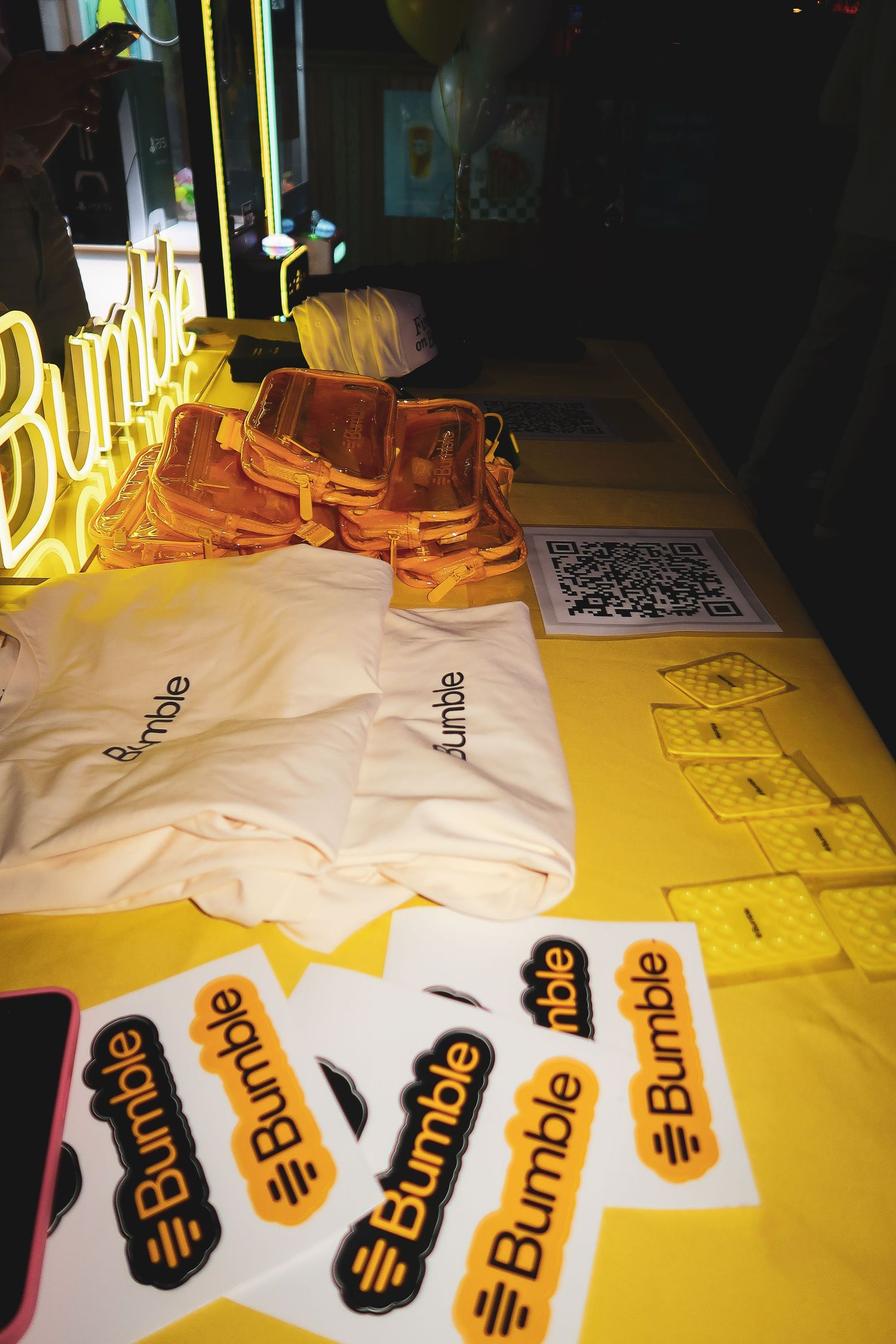 Yellow neon Bumble logo sign illuminates yellow table with stickers and branding materials laid out.