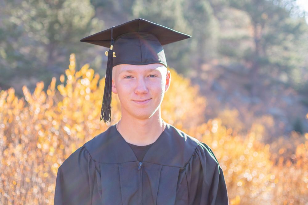 Graduate wearing black cap and gown stands smiling outdoors with golden autumn foliage in background.