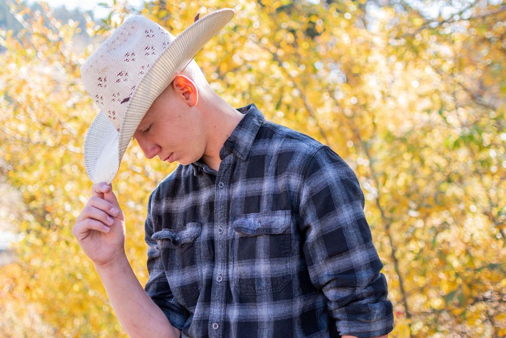 Person wearing plaid shirt and cowboy hat poses against backdrop of yellow autumn foliage.