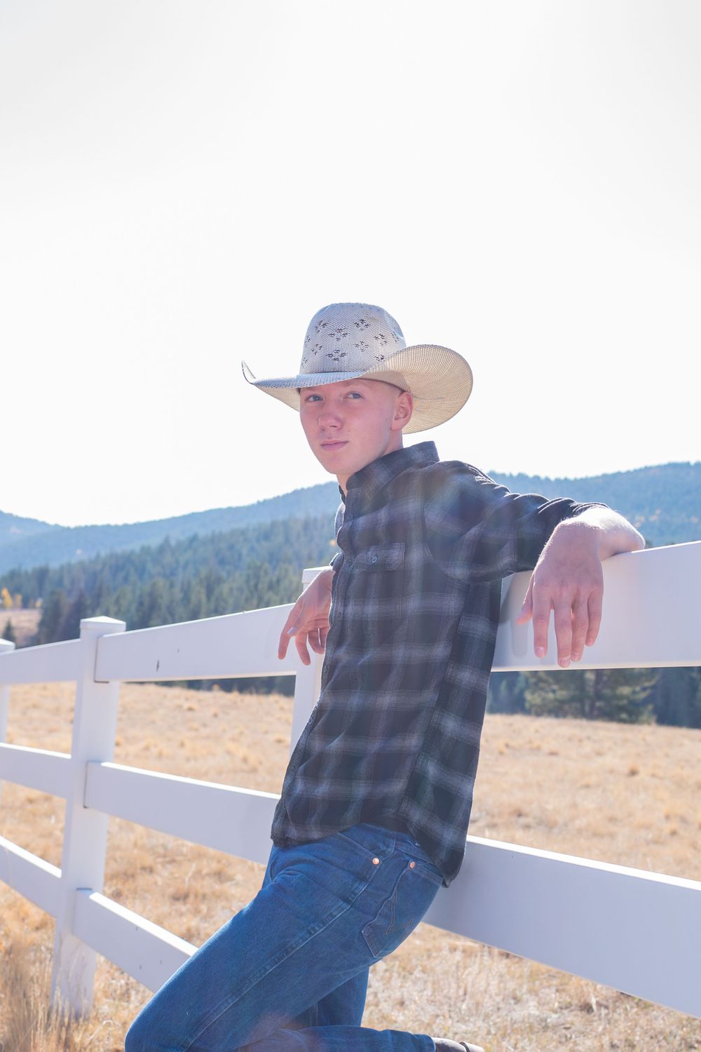 A person in a cowboy hat and plaid shirt leans against a white fence with mountains in the background.