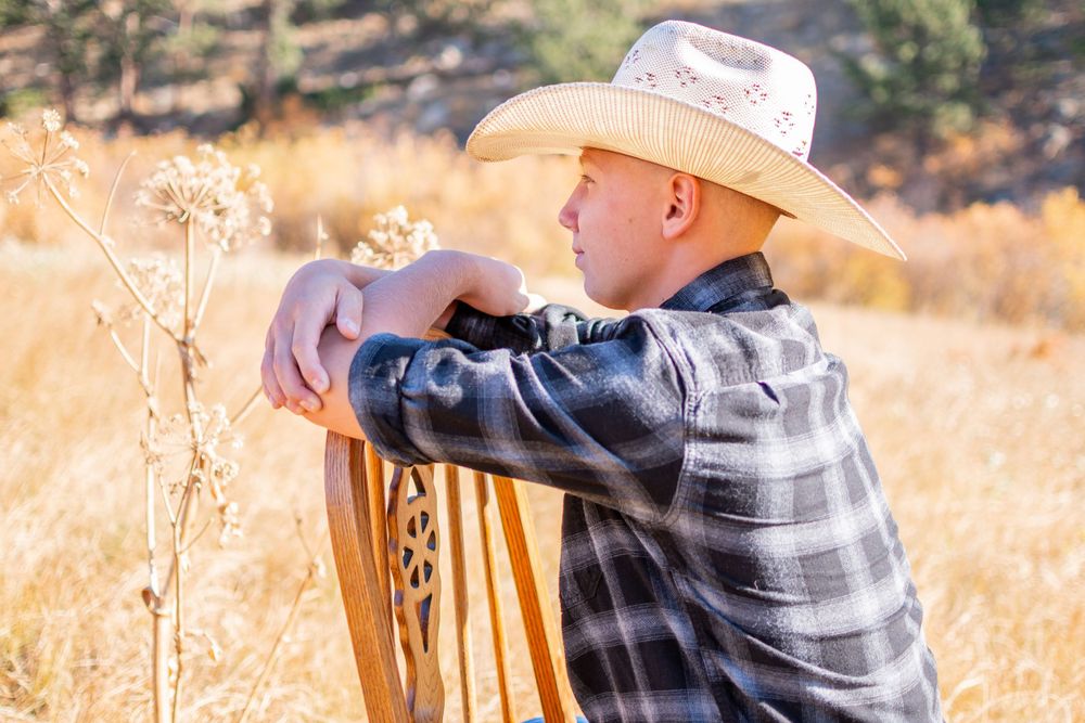 A person in a plaid shirt and cowboy hat leans on a wooden fence post in a sun-lit field.