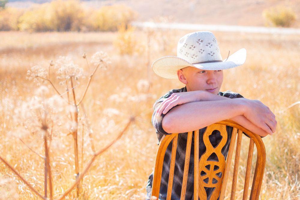 A person in a white cowboy hat sits backwards on a wooden chair amongst golden wheat fields in warm sunlight.