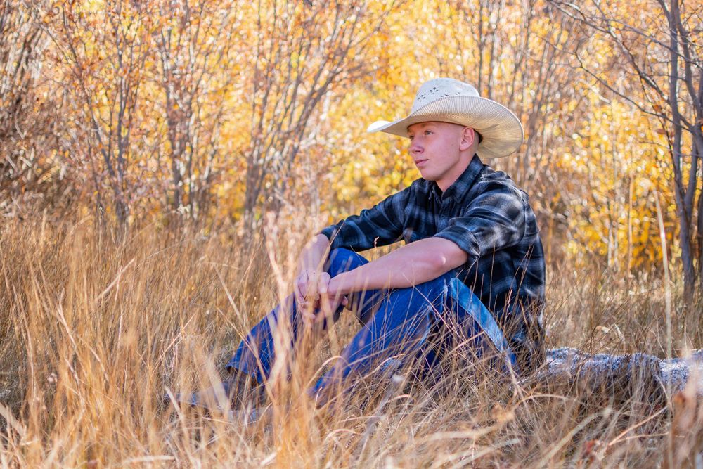 Person in cowboy hat and plaid shirt sits thoughtfully in golden autumn field during sunset.