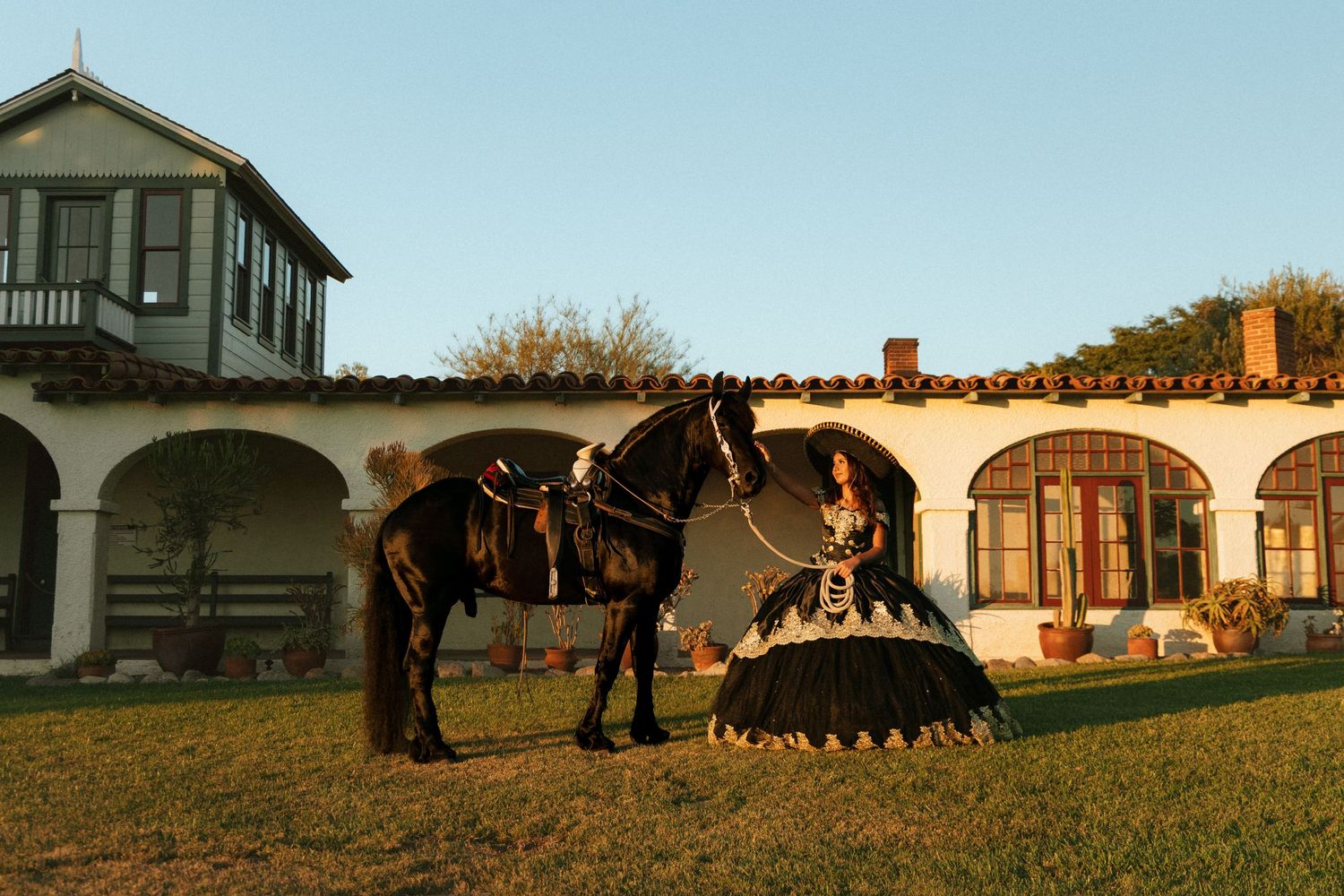 Black horse with saddle stands on grass in front of Spanish colonial style building at sunset.