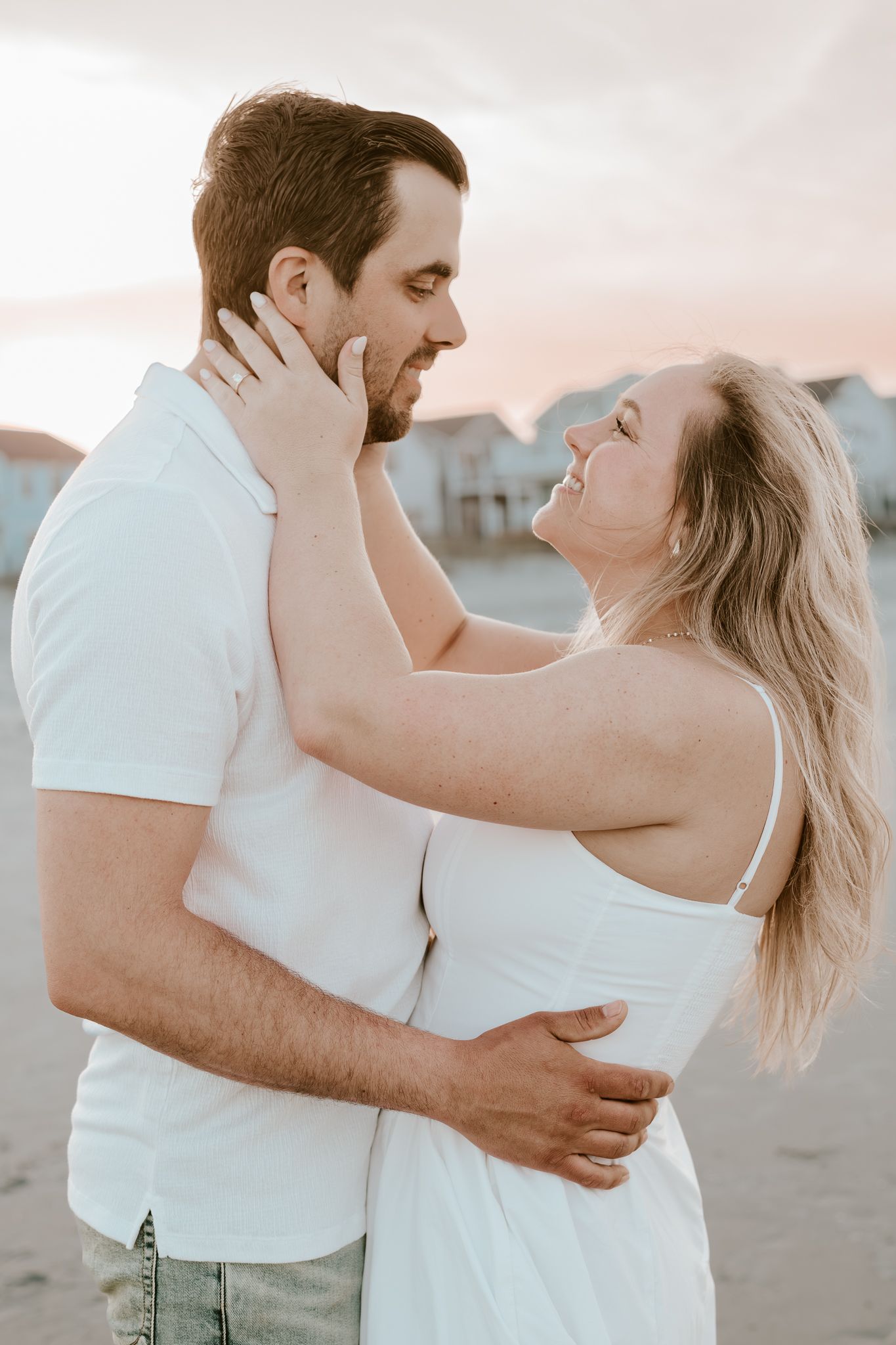 Romantic couple celebrates their engagement by embracing intimately on a beach wearing white against the soft sunlight.