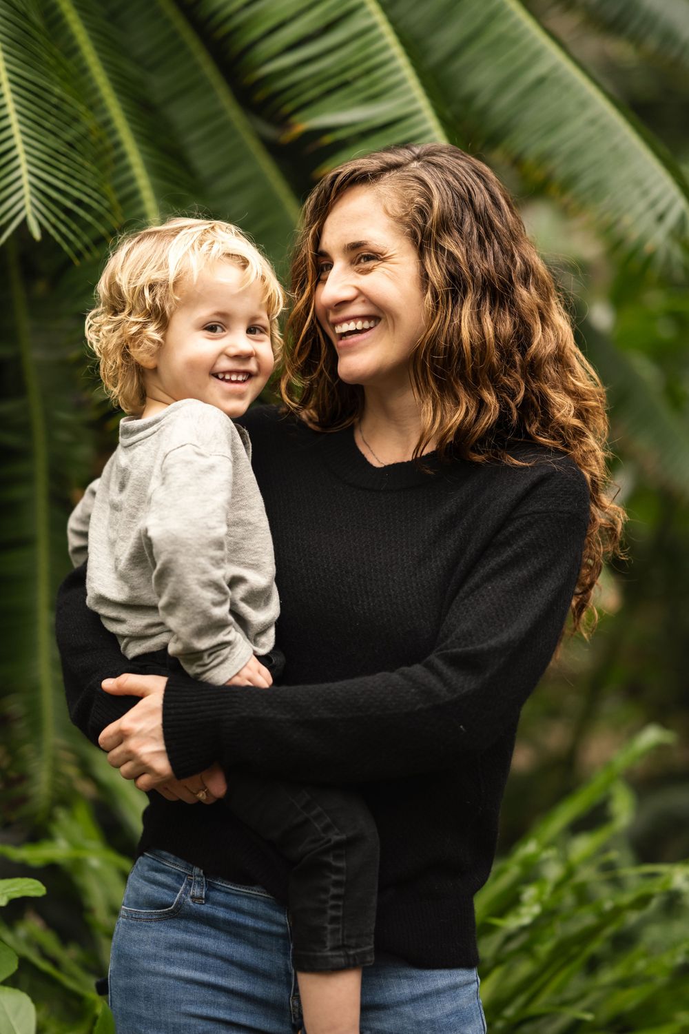 Mom and dad kissing while children peek through palm leaves – artistic family session in botanical garden – Ann Arbor photographer