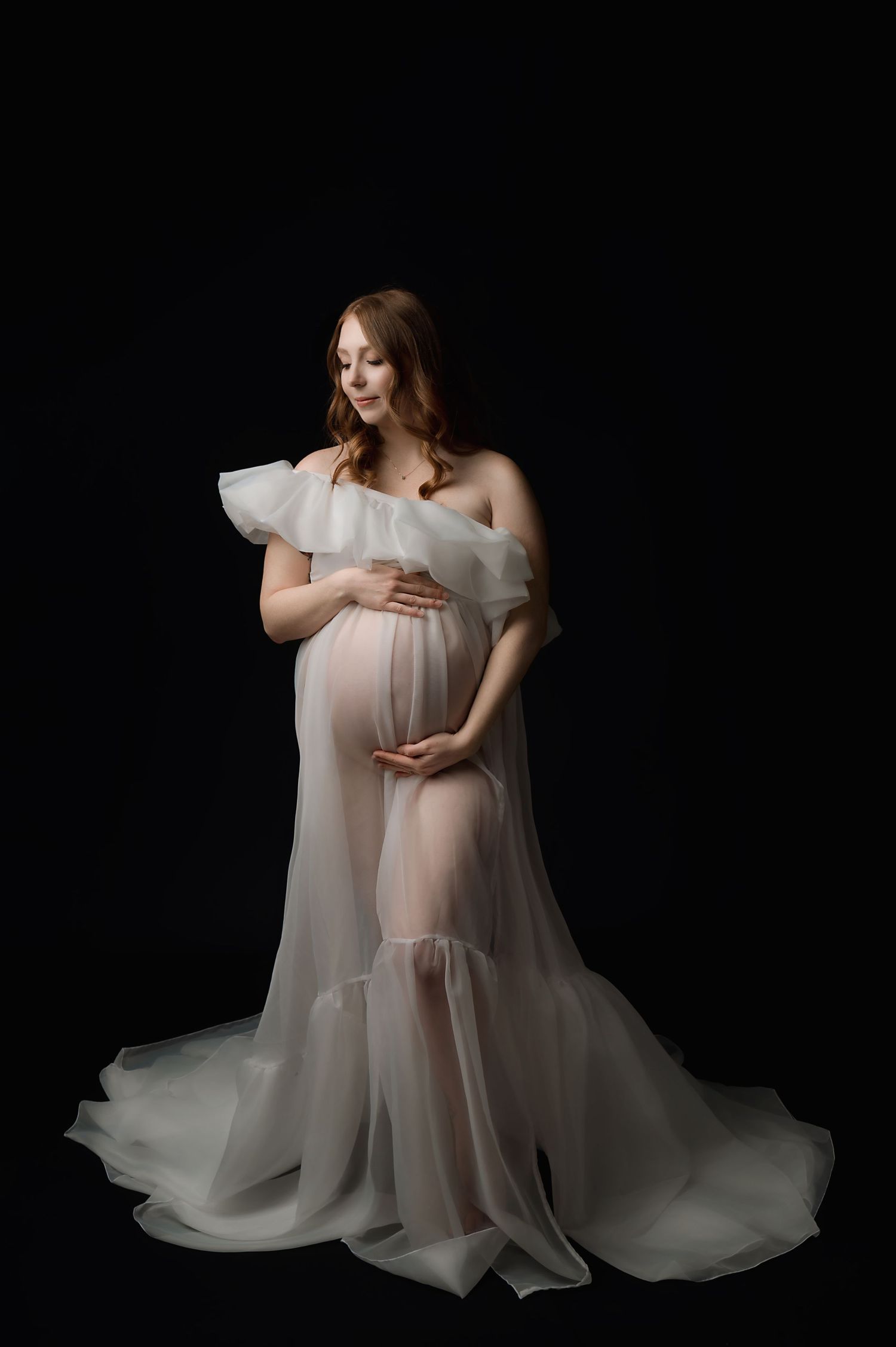 Ethereal maternity portrait featuring a flowing white sheer gown against a dark backdrop in dramatic studio lighting.