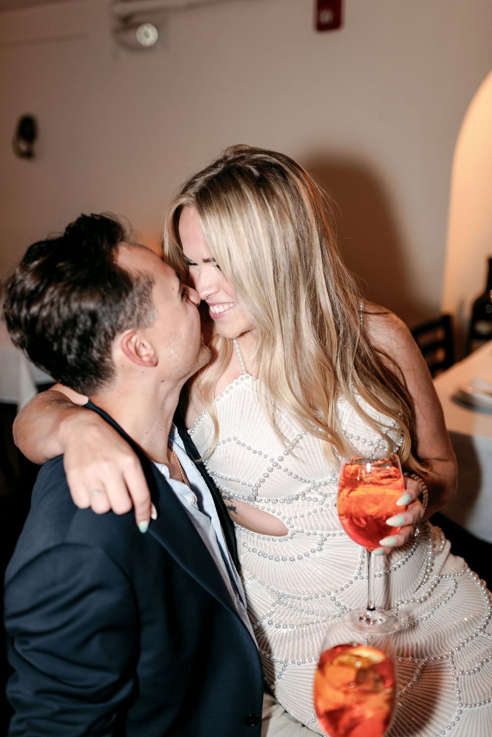 Couple sharing a romantic moment at a party, woman in white dress holding a cocktail, man in dark blazer.