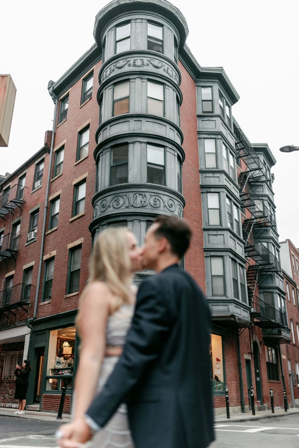 Couple sharing a kiss on a Boston street corner with a historic brick building featuring ornate bay windows in the background.
