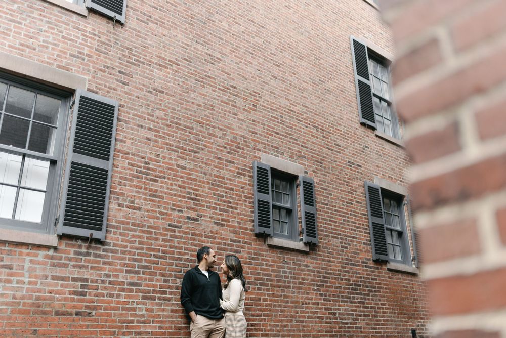 A couple stands close together against a rustic brick wall with black-shuttered windows in an urban setting.