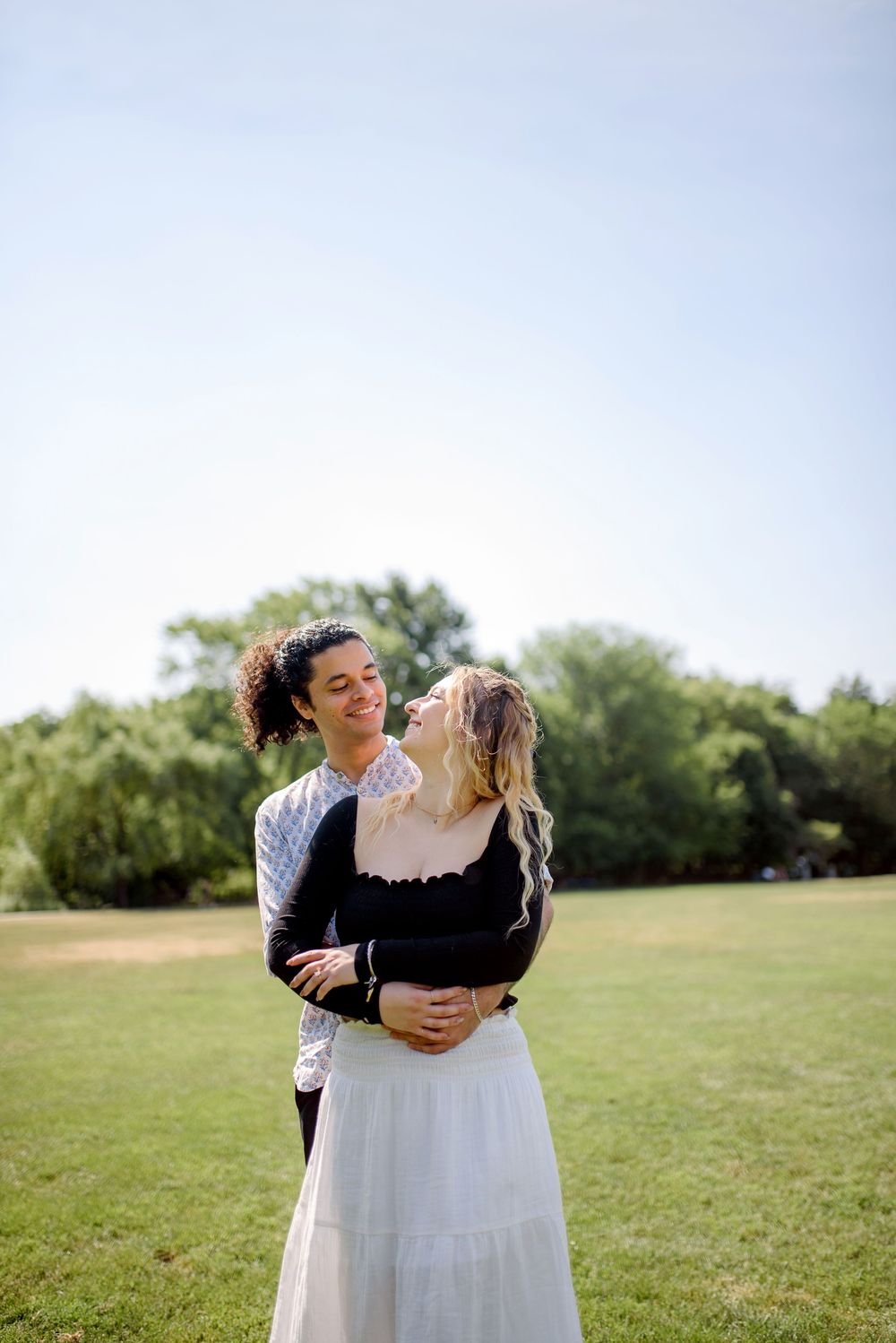 A couple embraces in a tender moment on a sunlit grassy field with trees in the background.