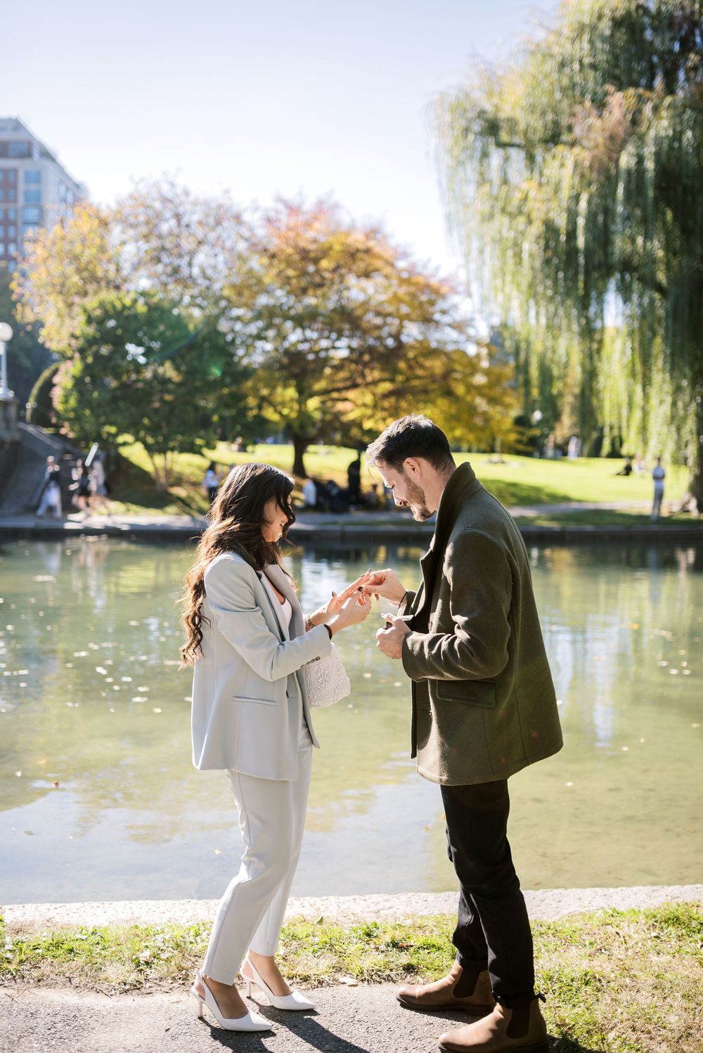 Couple examines ring by pond in Boston Public Garden surrounded by fall foliage.