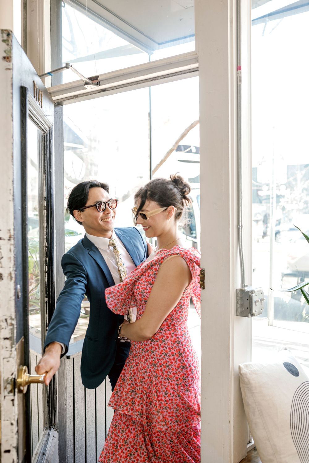 A couple in formal attire share a romantic moment by a bright window, with her in a red floral dress and him in a navy suit.