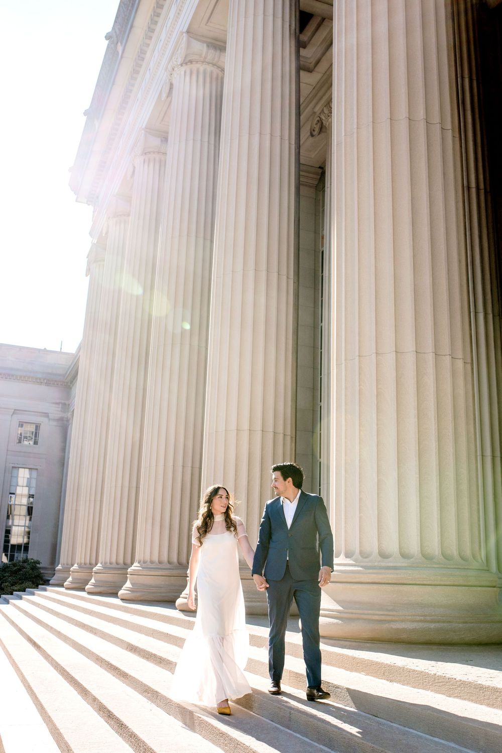 A couple in wedding attire stands between grand white columns casting dramatic shadows on courthouse steps.