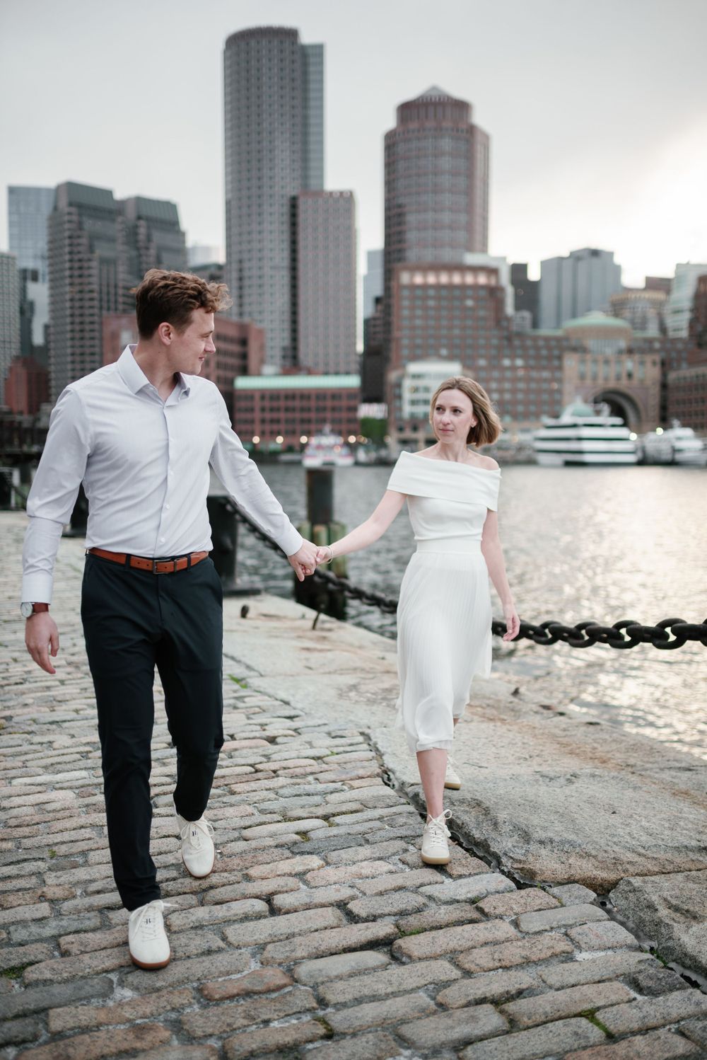 Couple holds hands while walking along cobblestone path with Boston Harbor and downtown buildings behind them.