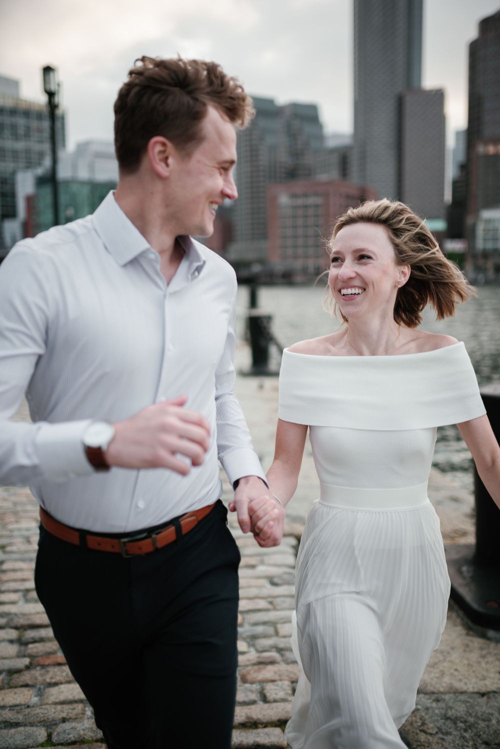 Joyful couple runs along cobblestone path by Boston Harbor during engagement photoshoot.