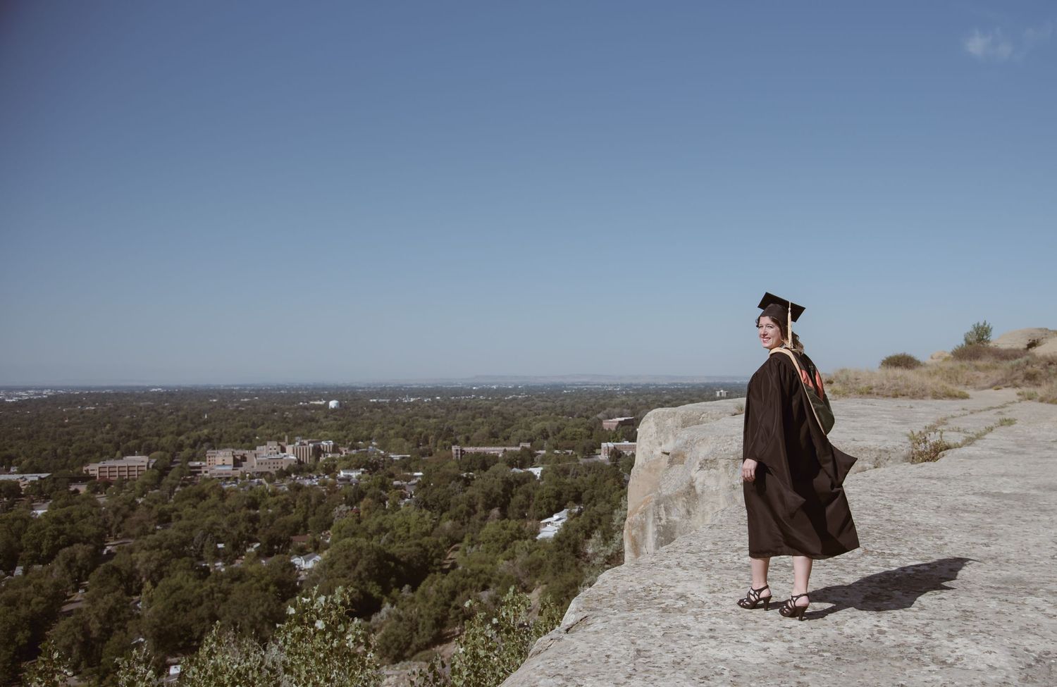 Female graduate in cap and gown posing on a cliff edge overlooking a landscape. Whidbey Island Senior Graduation portrait