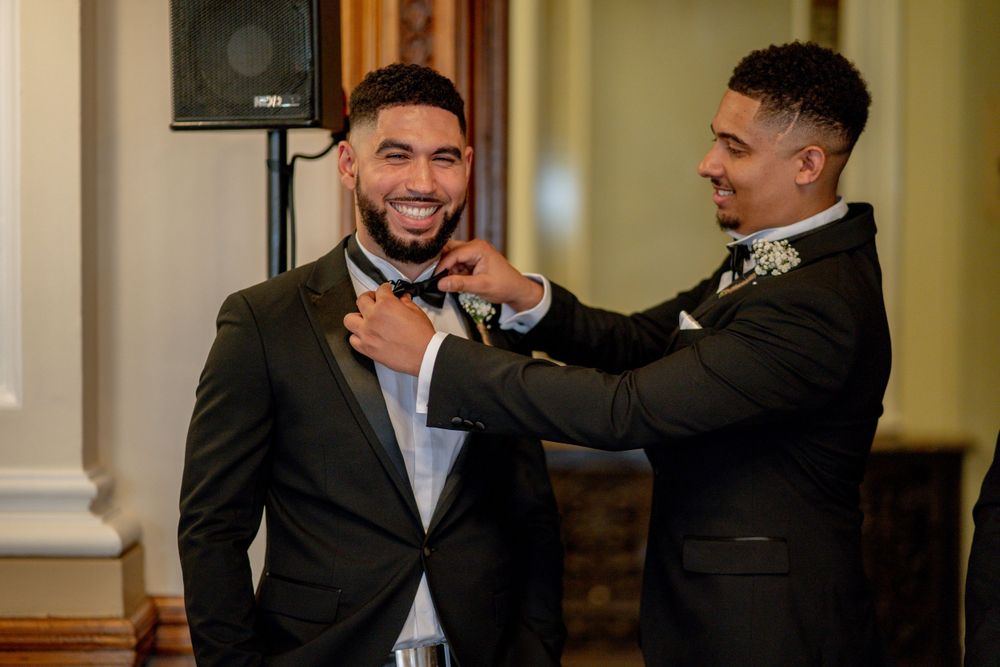 Best man helps adjust the groom's tie and jacket before the wedding ceremony in an elegant indoor setting.