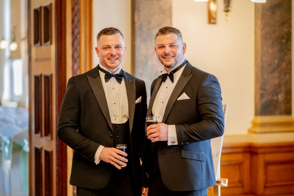Two men in matching black tuxedos and bow ties smile while holding drinks in an elegant venue.