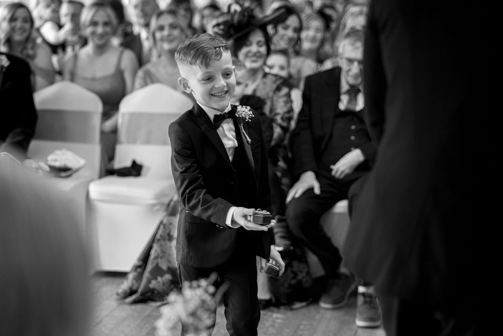 Young ring bearer in a tuxedo walks down the wedding aisle during ceremony while guests watch in black and white photo.