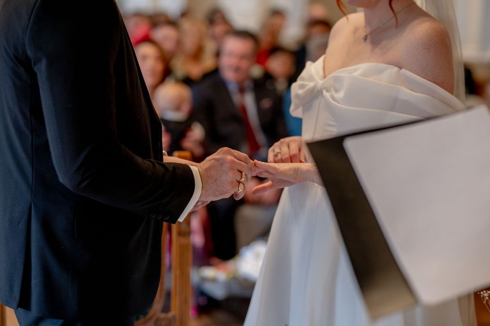 Close-up shot of exchanging wedding rings during ceremony with guests watching in background.