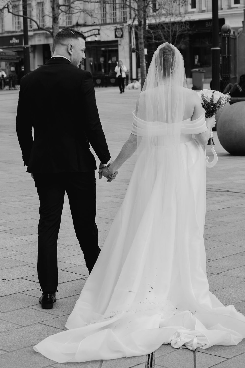 A black and white photo of a bride in a flowing white dress and groom in dark suit walking hand in hand on a city street.