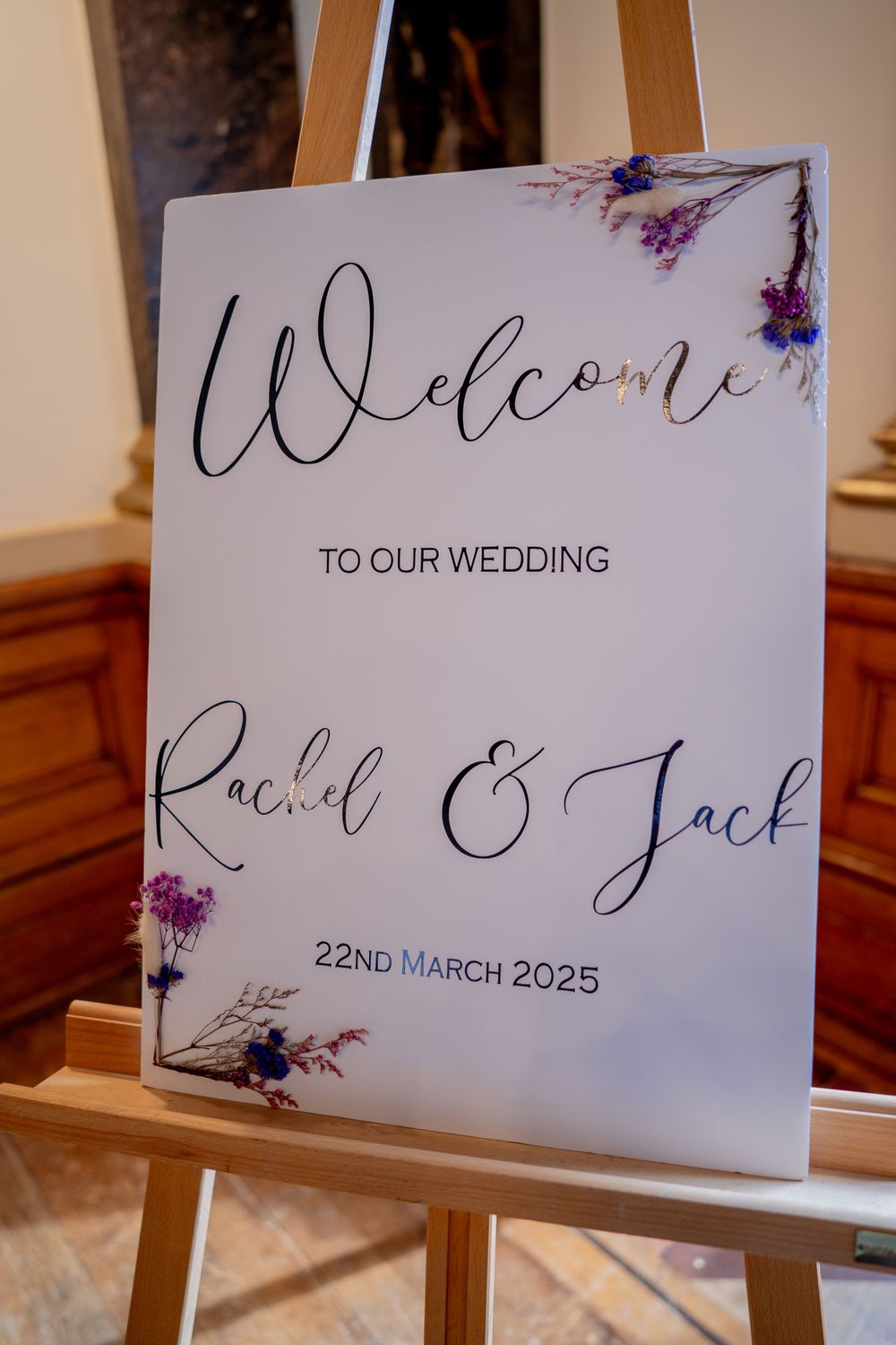 Elegant wedding welcome sign with floral details and March 2025 date displayed on wooden easel.