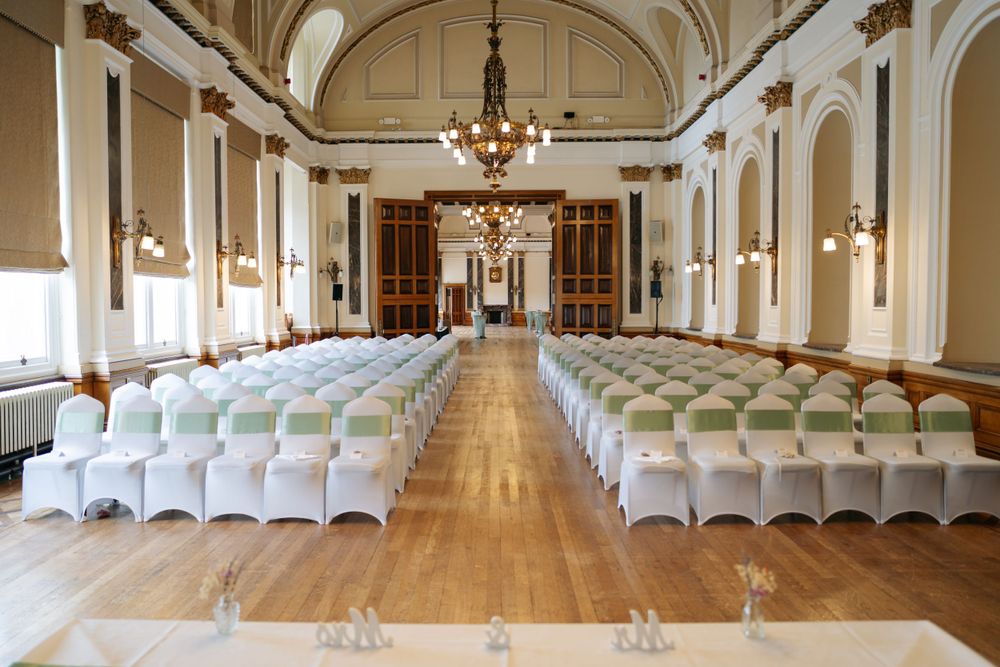 Elegant ballroom with white chairs in rows, crystal chandeliers, and ornate architectural details set for a formal ceremony.