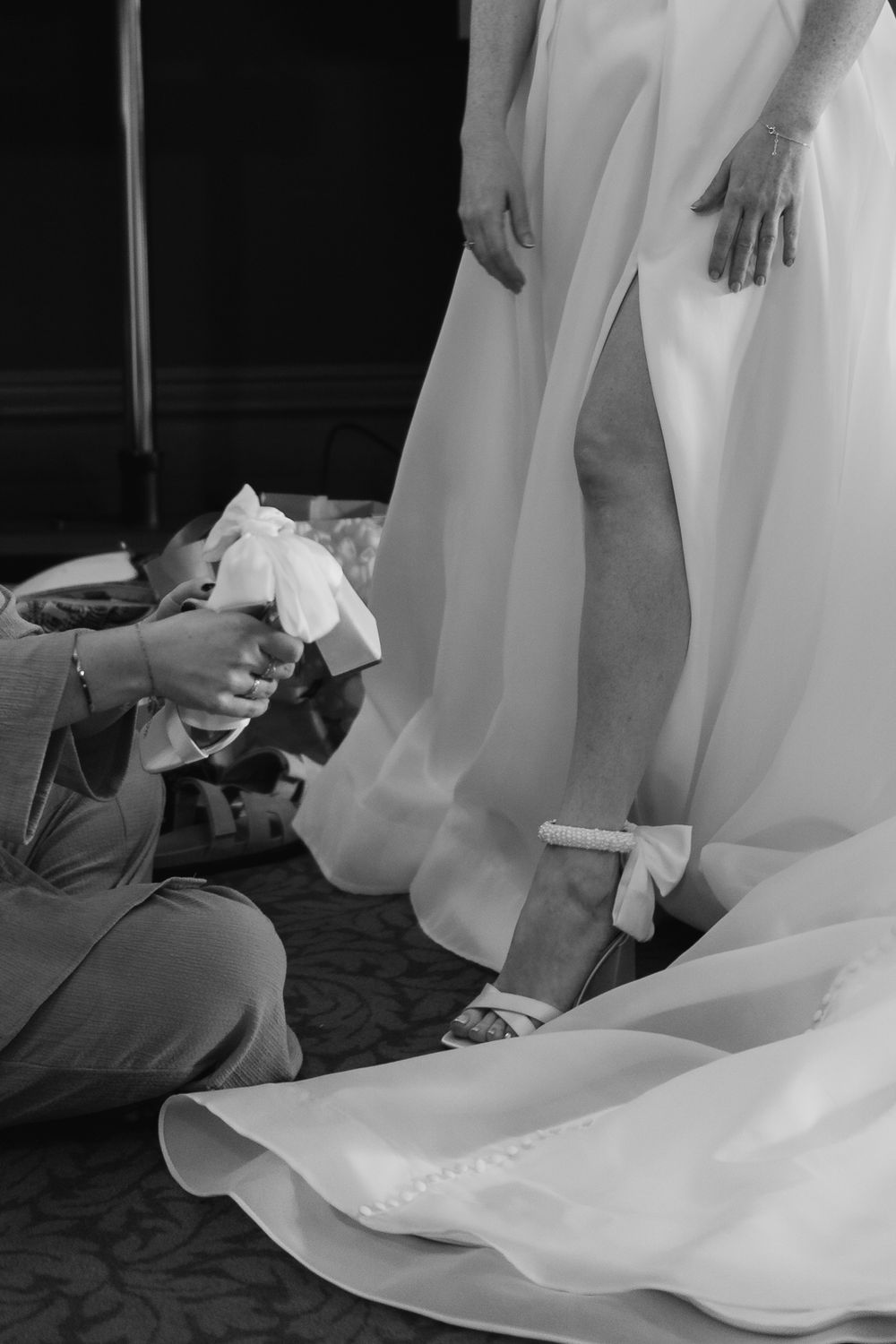 Black and white photo of someone helping to adjust the hem and shoes of a flowing white wedding dress.