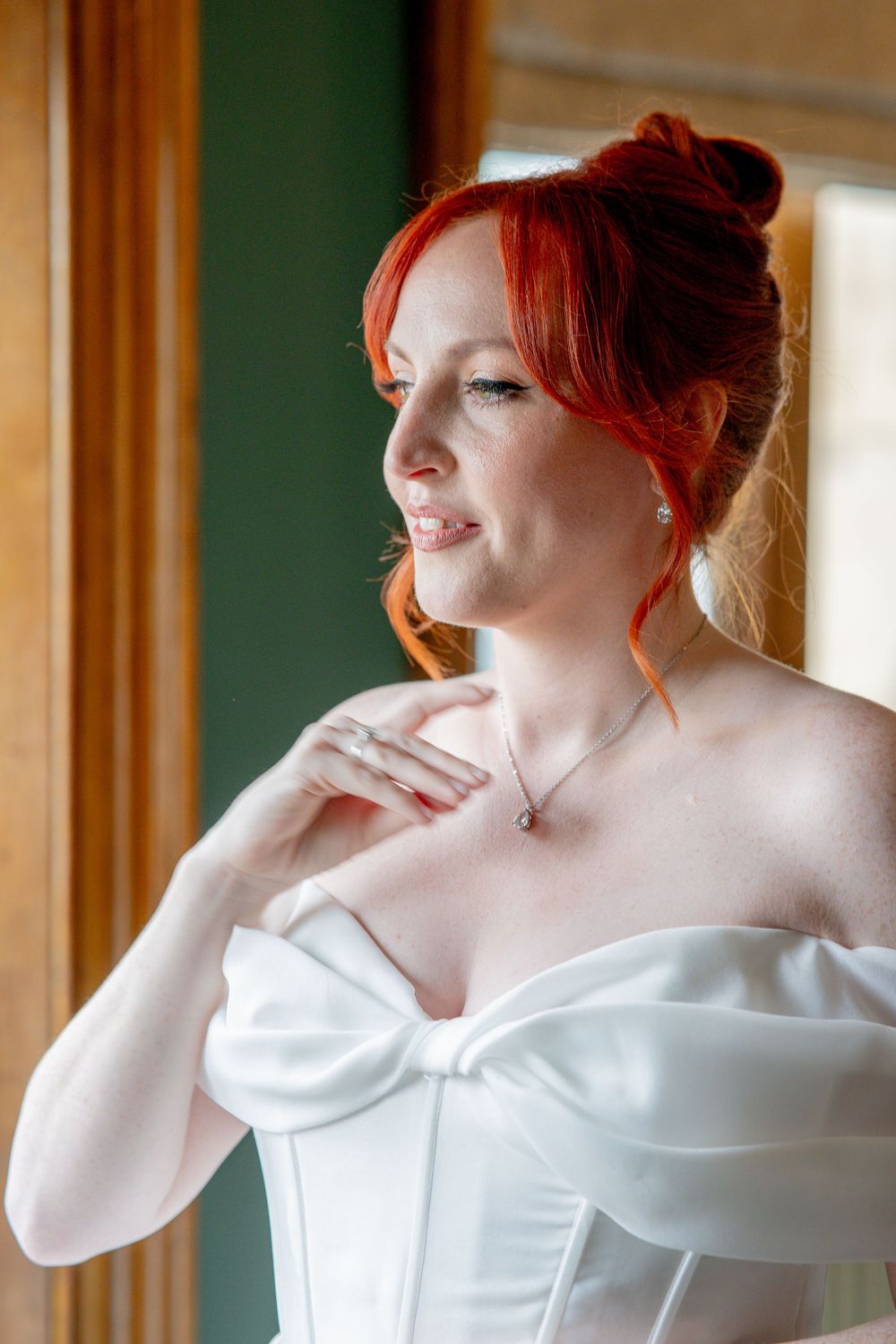 Portrait of someone in a white corset wedding dress with vibrant red hair styled up, gazing thoughtfully by a window.