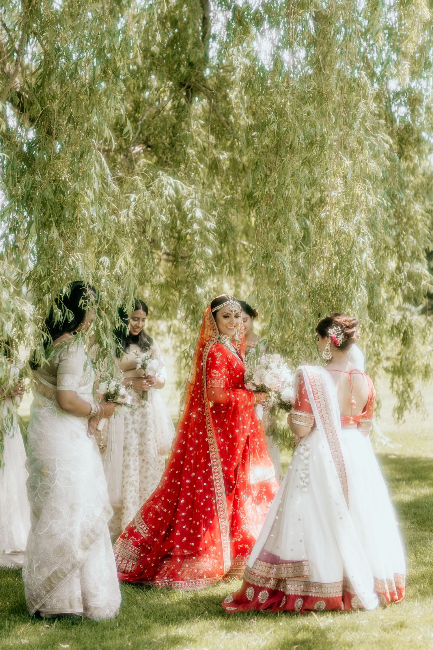 A group in traditional South Asian wedding attire stands beneath willow trees in a dreamy ethereal outdoor setting.