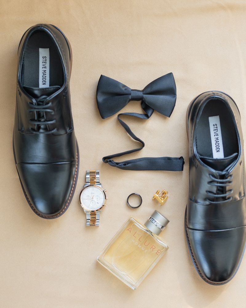 Black dress shoes arranged with a bow tie, watch, rings, and cologne bottle in a groom's accessories flatlay.