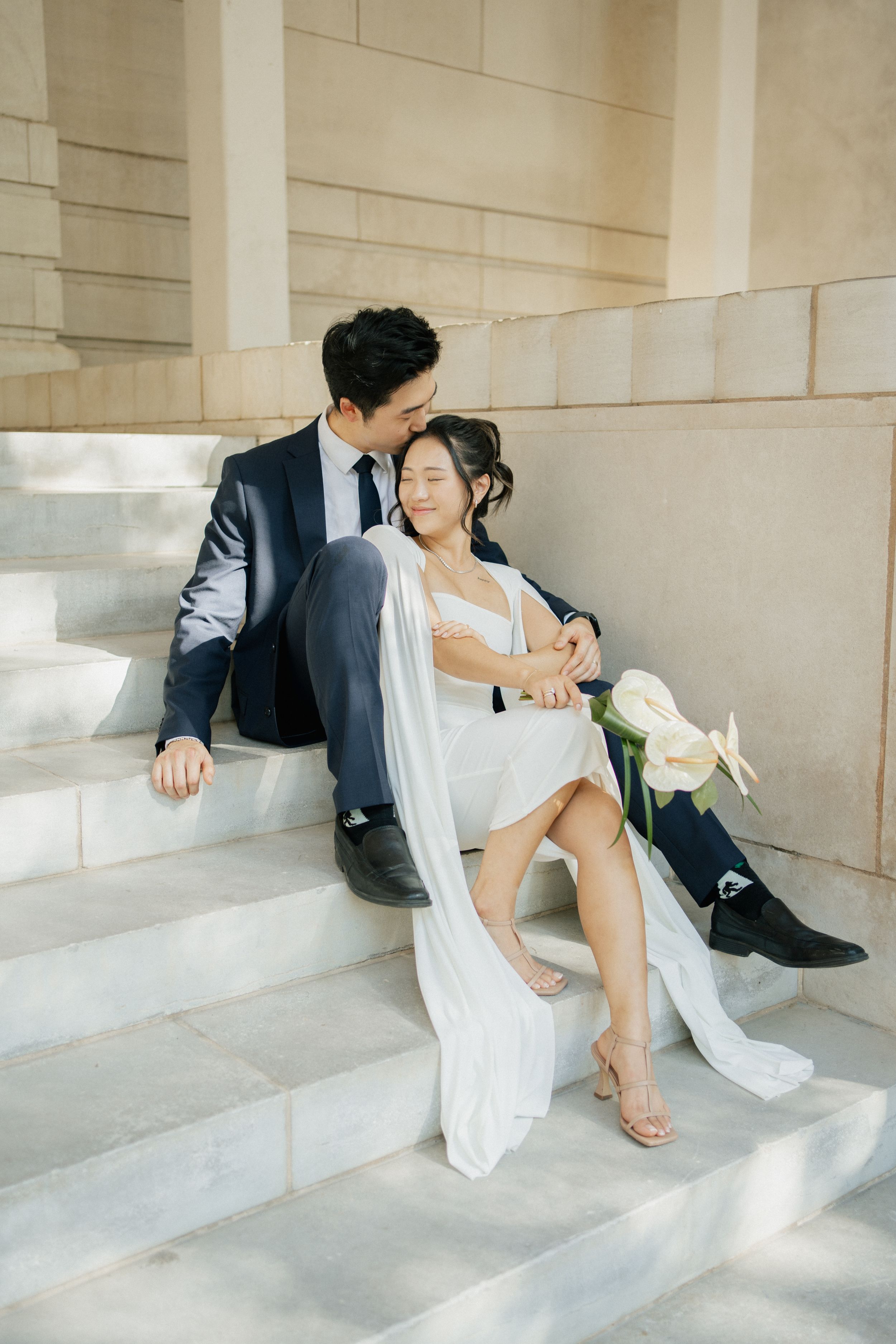 Newlywed couple rests elegantly on stone steps with a flowing white veil and calla lily bouquet in natural light.
