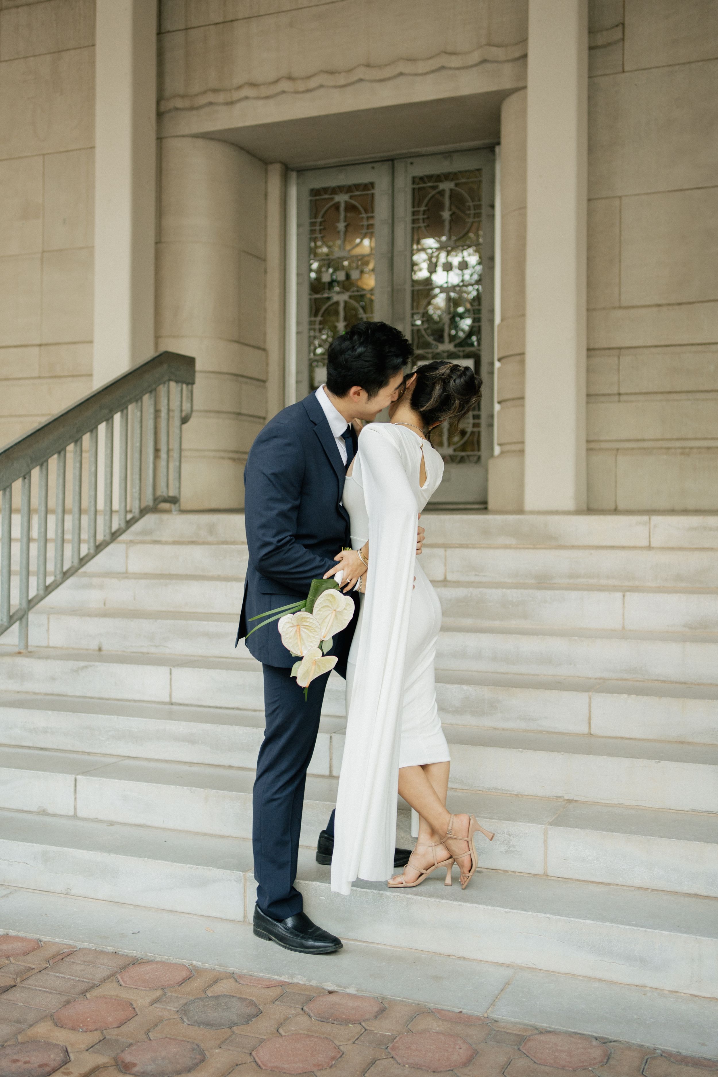 A couple sharing a tender moment on stone steps outside an elegant building entrance with white pillars.