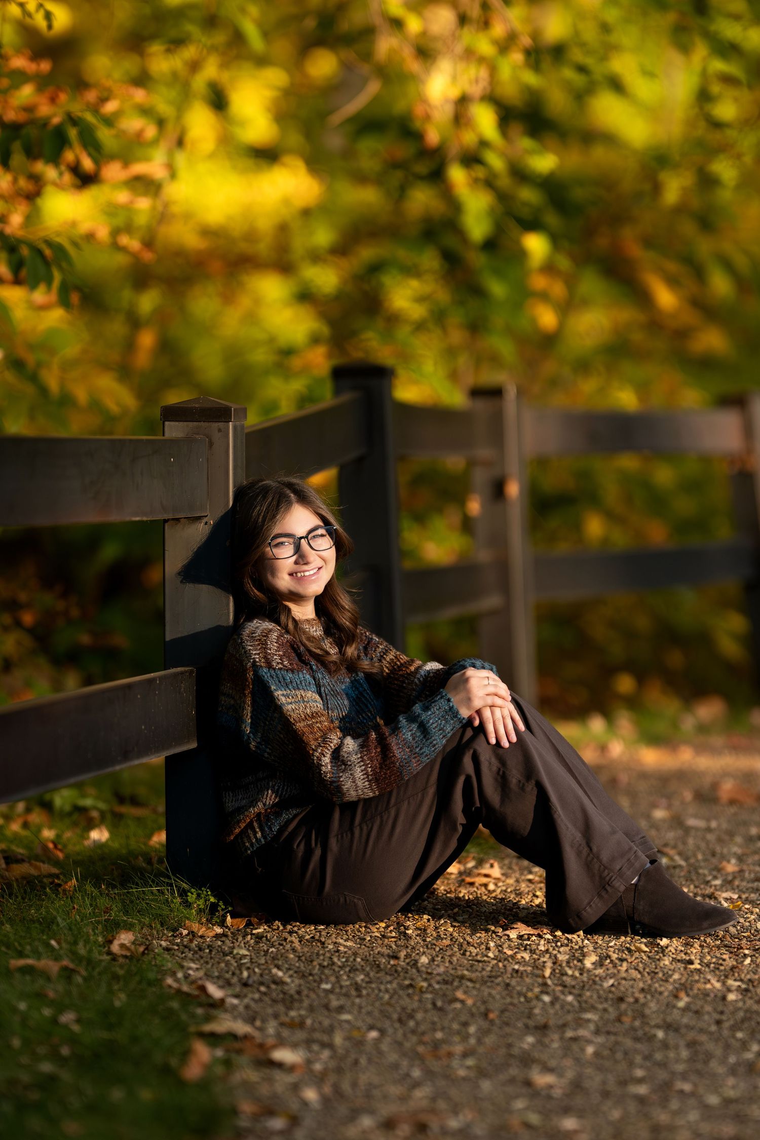Young woman with glasses sitting against a wooden fence surrounded by vibrant autumn foliage in golden light.