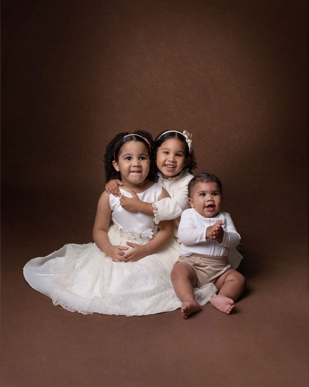 Three siblings wearing white formal attire pose together for a portrait against a brown background.