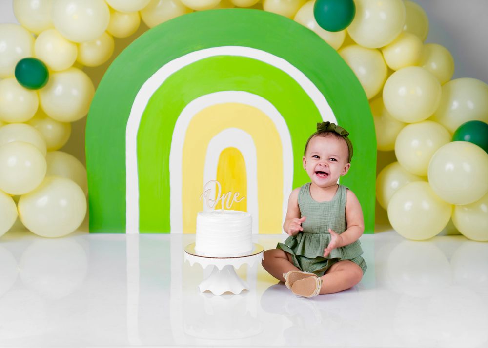 Rainbow backdrop with yellow balloons and green arch for baby photography.