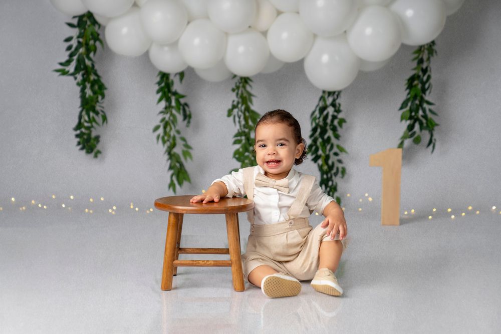 Smiling baby celebrates milestone under white balloon arch and greenery decorations.