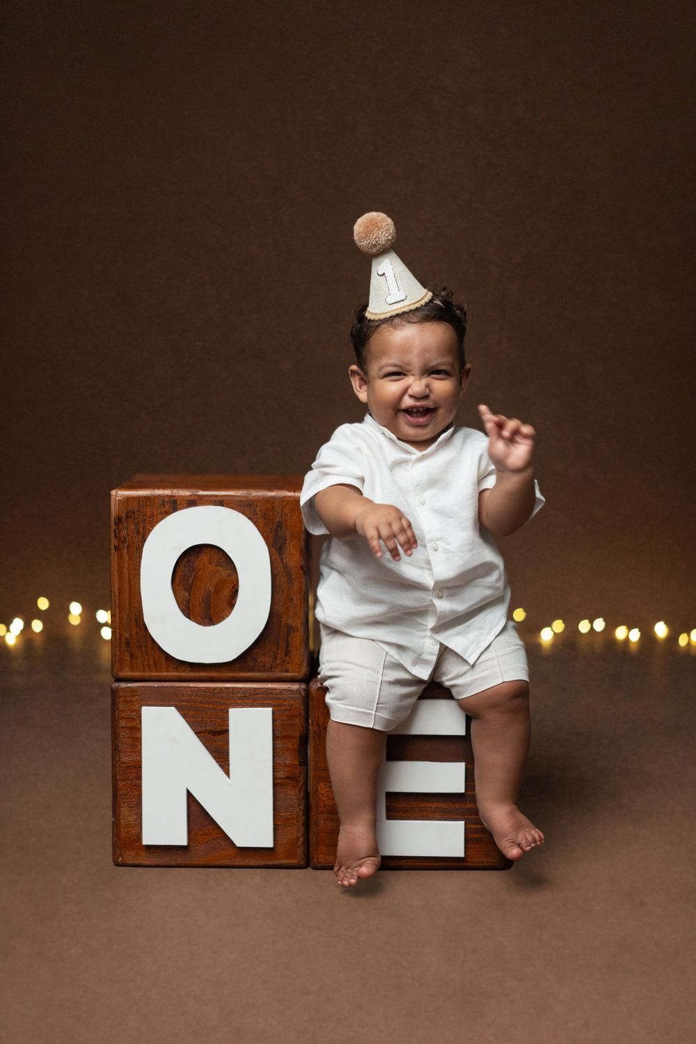 Joyful baby wearing party hat sits on wooden blocks spelling 'ONE' surrounded by twinkling lights.