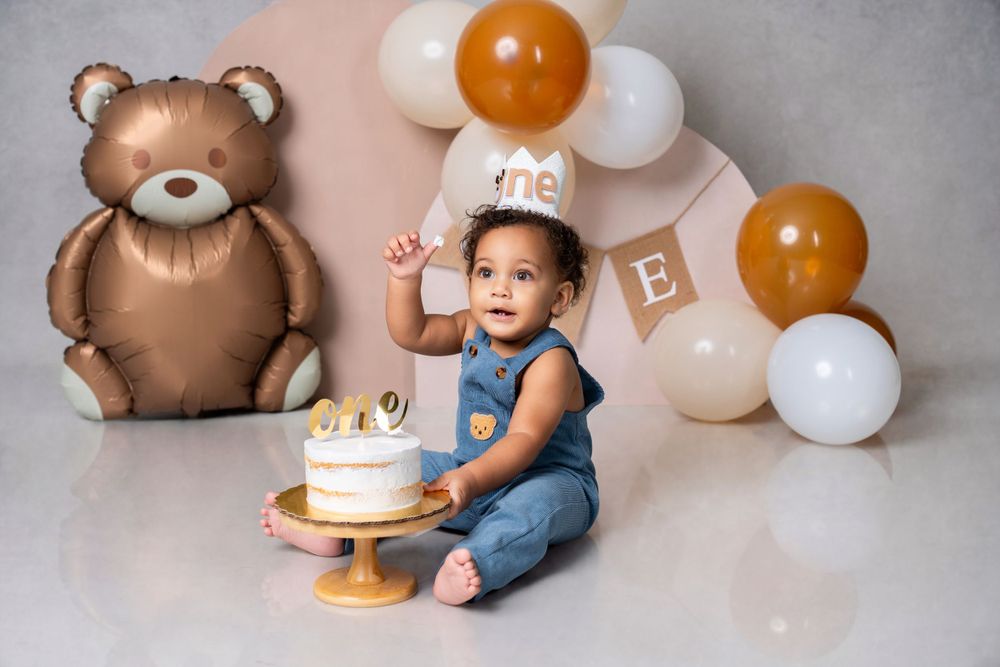 Baby celebrating first birthday with cake next to large teddy bear and colorful balloons.