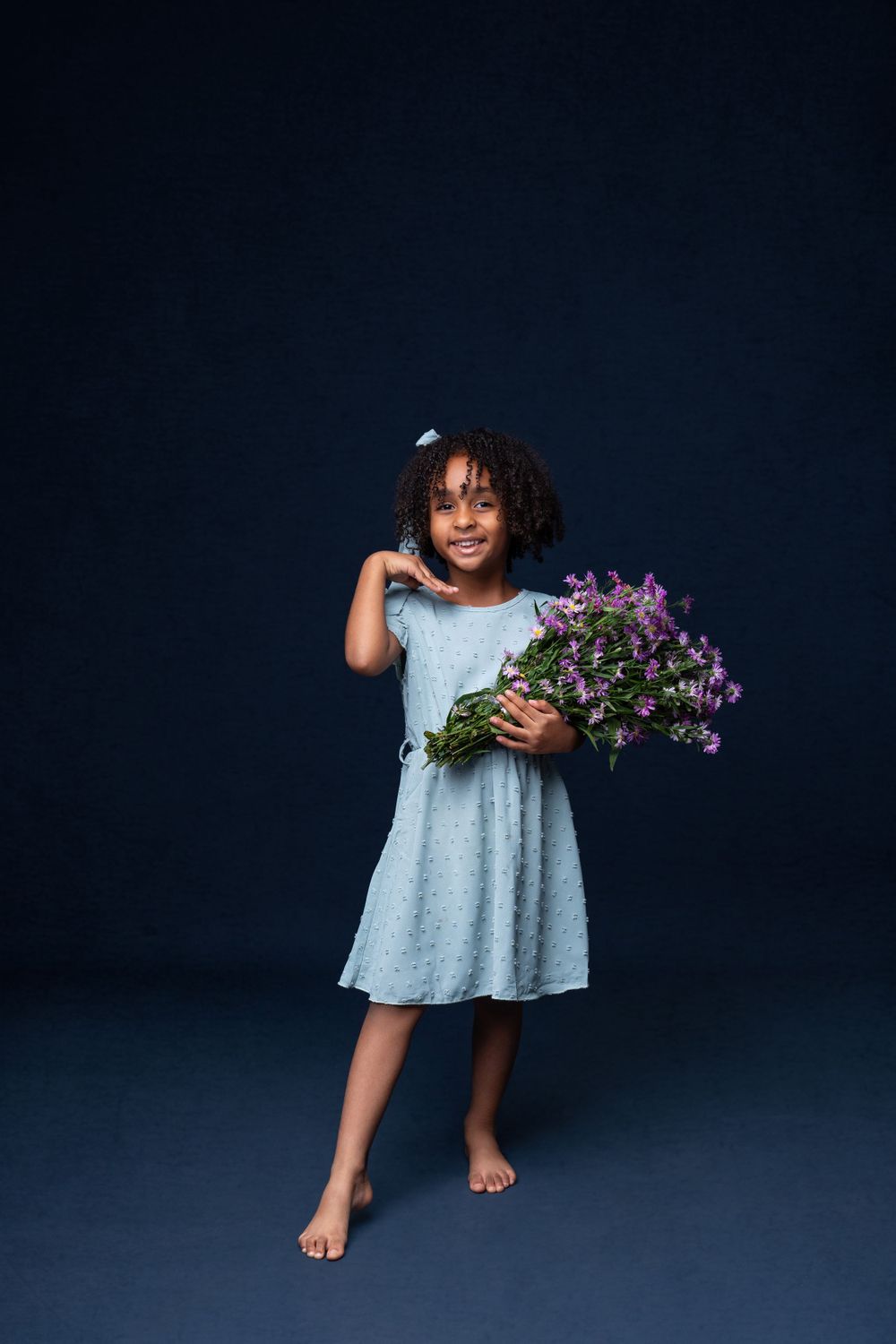 Young barefoot child in blue dress holds purple wildflowers against dark background.